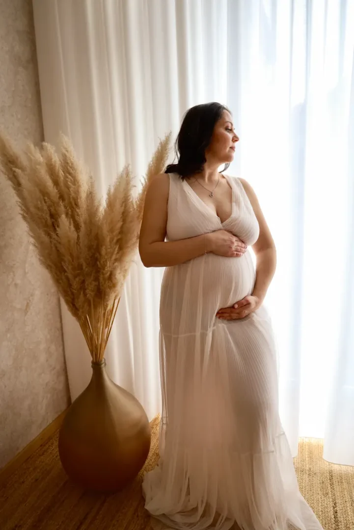 Schwangere im weißen Kleid steht am hellen Fenster neben einer Vase mit Pampasgras