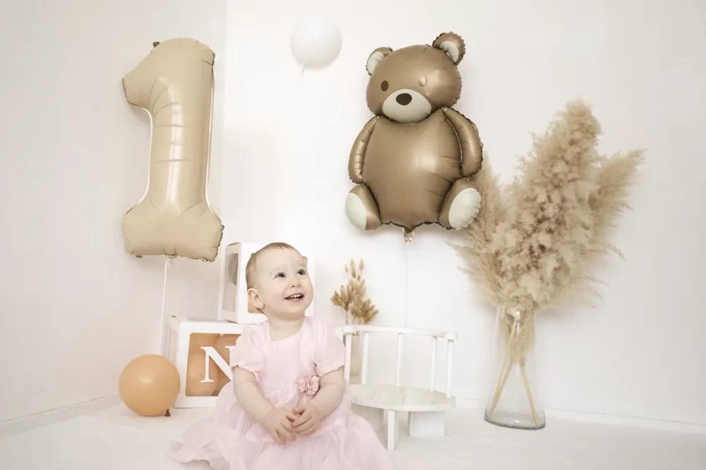 Kleinkind im rosa Kleid sitzt zwischen Ballons, einer Eins und einem Teddyballon