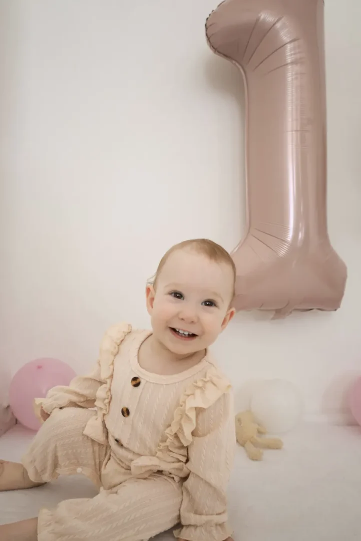 Kleinkind sitzt lachend vor einem großen rosa Ballon in Form einer Eins