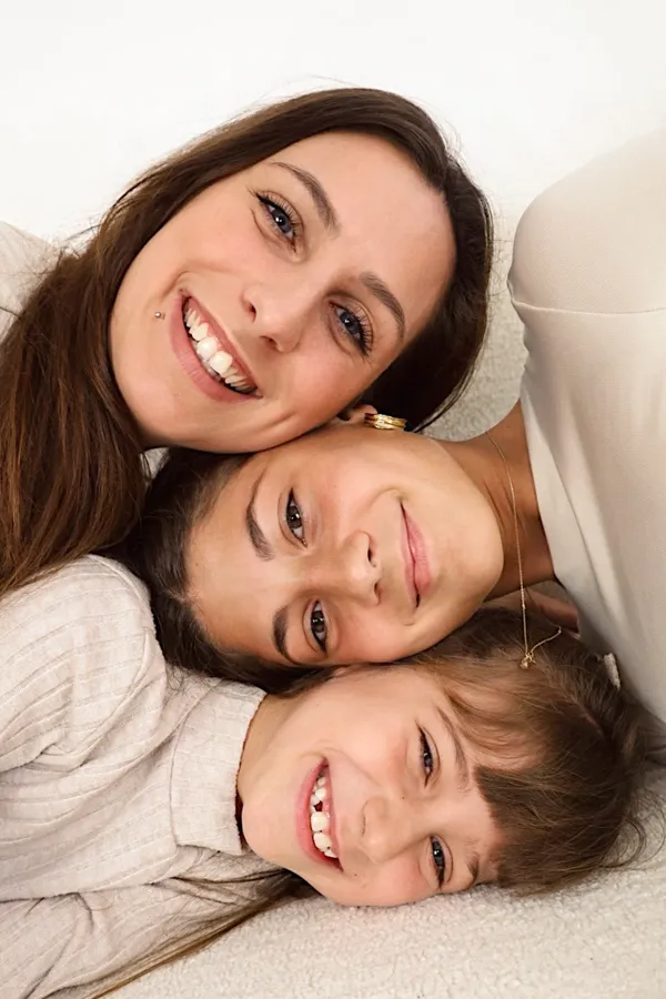 Mutter und zwei Töchter liegen Kopf an Kopf auf dem Sofa und lachen in die Kamera