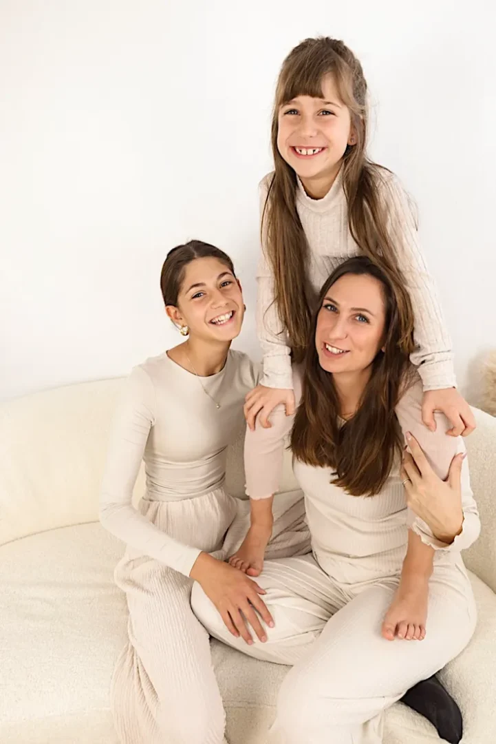Mutter und zwei Töchter in hellen Outfits lachen gemeinsam auf einem weißen Sofa