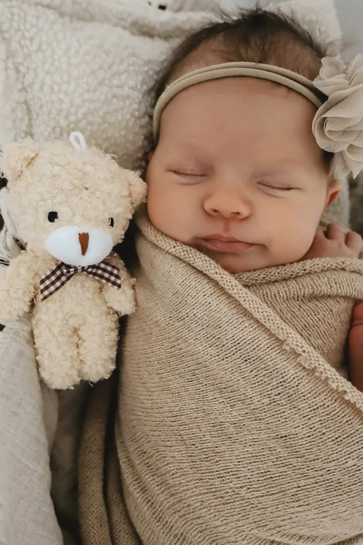 Neugeborenes schläft mit Haarband in eine beige Strickdecke gewickelt, daneben ein Teddy